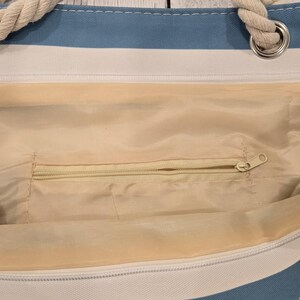 May include: A blue and white striped tote bag with rope handles. The interior of the bag is a light beige color and features a zippered pocket. The bag is designed for carrying items and has a casual, beachy aesthetic.