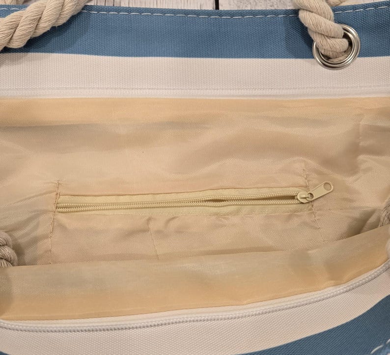 May include: A blue and white striped tote bag with rope handles. The interior of the bag is a light beige color and features a zippered pocket. The bag is designed for carrying items and has a casual, beachy aesthetic.