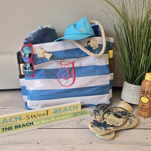 May include: A blue and white striped beach tote bag with a pink embroidered initial. The bag is filled with a blue towel, sunglasses, and a blue cap. A wooden sign reads "Beach Sweet Beach". Also shown are flip-flops and sunscreen.