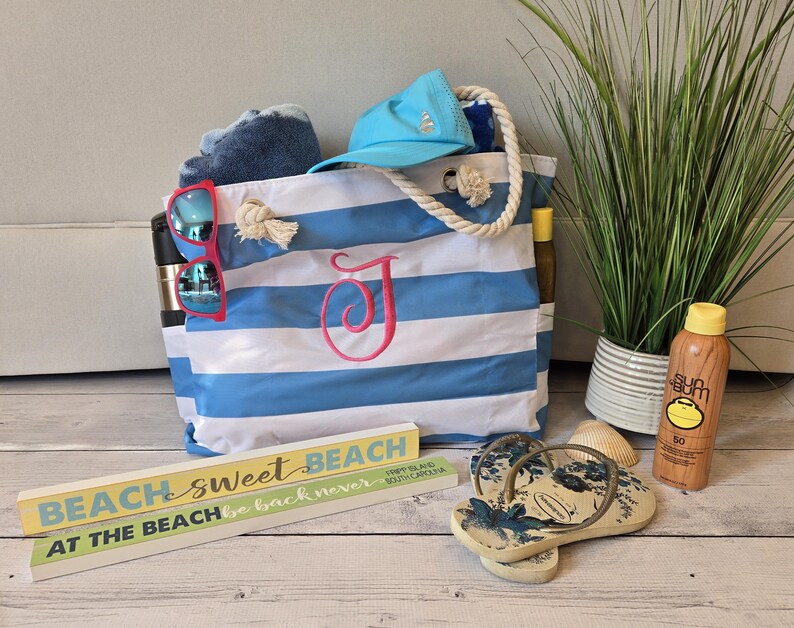 May include: A blue and white striped beach tote bag with a pink embroidered initial. The bag is filled with a blue towel, sunglasses, and a blue cap. A wooden sign reads "Beach Sweet Beach". Also shown are flip-flops and sunscreen.