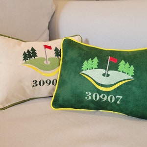 May include: Two decorative pillows with a golf course design. The pillows are green and beige with yellow trim. The green pillow has the number 30907 embroidered on it. The beige pillow has the number 3090 embroidered on it.