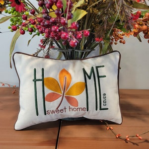 May include: A white pillow with a brown border features an embroidered design of a stylized leaf in shades of orange, yellow, and brown. The words "HOME" and "sweet home" are embroidered in green, and the number "63123" is embroidered in black.
