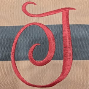 May include: Close-up of a fabric with a pink embroidered letter "J". The background features alternating horizontal stripes of light blue and off-white. The embroidery thread has a textured appearance.