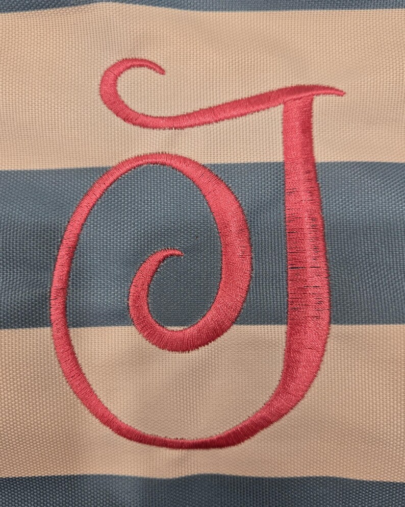 May include: Close-up of a fabric with a pink embroidered letter "J". The background features alternating horizontal stripes of light blue and off-white. The embroidery thread has a textured appearance.