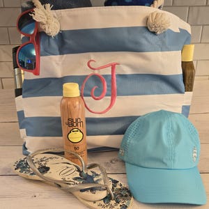 May include: A beach-themed collection featuring a blue and white striped tote bag with a pink embroidered "J", sunglasses, sunscreen, flip-flops, and a light blue cap. The bag is filled with towels.