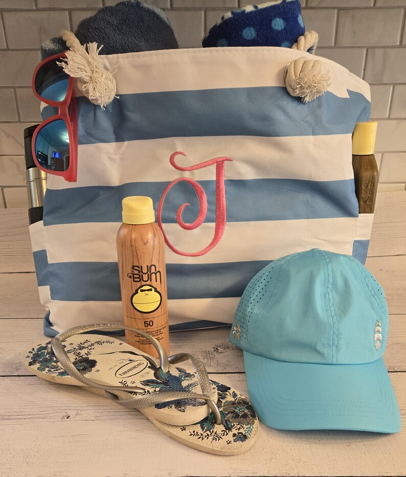 May include: A beach-themed collection featuring a blue and white striped tote bag with a pink embroidered "J", sunglasses, sunscreen, flip-flops, and a light blue cap. The bag is filled with towels.