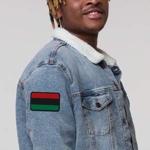 Puede incluir: Chaqueta vaquera azul claro con cuello de borreguillo blanco. La chaqueta presenta un parche rectangular en el brazo izquierdo con un diseño negro, rojo y verde. Tiene dos bolsillos en el pecho y cierre de botones.