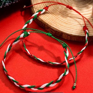 Könnte beinhalten: Zwei geflochtene Armbänder mit roten, weißen und grünen Fäden, vor einem leuchtend roten Hintergrund. Die Armbänder sind verstellbar und haben goldfarbene Perlen. Eines liegt auf Holz.