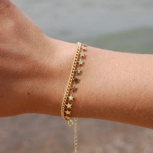 May include: A gold-colored bracelet with a chain and a row of small star charms. The bracelet is worn on a wrist, with a blurred background of water. The jewelry is delicate and likely made of metal.