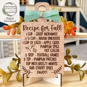 May include: A wooden sign shaped like a cutting board with the text "Recipe for Fall" and a list of ingredients for a fall recipe. The ingredients include crisp mornings, warm sweaters, apple cider, pumpkin spice, hot cocoa, football games, pumpkin patches, breezy nights, and starry skies. The sign is decorated with a green bow and is sitting on a wooden stand.