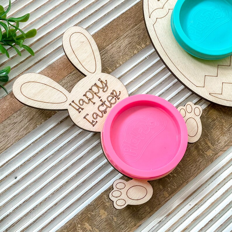 May include: Wooden bunny-shaped Easter craft with the words "Happy Easter" engraved on the side. The bunny has a pink play dough container in the center.