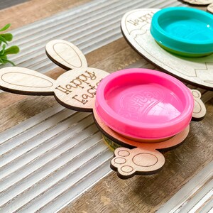May include: Two wooden Easter-themed play dough holders. One is shaped like a bunny with the text "Happy Easter" and the other is shaped like an egg. Both holders have a pink and a blue play dough container inside.