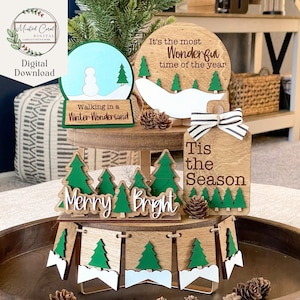 May include: A tiered wooden tray decorated with Christmas-themed wooden ornaments. The ornaments feature green trees, snowmen, and phrases like "It's the most Wonderful time of the year", "Walking in a Winter Wonderland", "Merry Bright", and "Tis the Season".