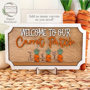 Pode incluir: Uma placa de madeira com moldura branca que diz "Bem-vindos ao nosso canteiro de cenouras". A placa tem recortes de cenouras laranja com topos verdes e nomes escritos abaixo de cada cenoura. Os nomes são Pai, Mãe e Joell.