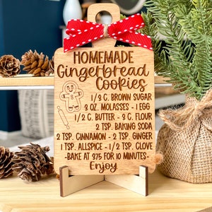 May include: Wooden gingerbread cookie recipe ornament with a red ribbon. The recipe includes 1/3 cup brown sugar, 3 ounces molasses, 1 egg, 1/2 cup butter, 3 cups flour, 2 teaspoons baking soda, 2 teaspoons cinnamon, 2 teaspoons ginger, 1 teaspoon allspice, 1/2 teaspoon clove. Bake at 375 degrees Fahrenheit for 10 minutes. Enjoy!