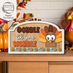 May include: Thanksgiving-themed wooden sign with the words "GOBBLE til you WOBBLE" in brown and orange lettering. A cartoon turkey is featured on the right. The sign has a light blue border with turkey leg and leaf accents.