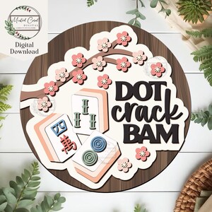 May include: A wooden sign featuring a decorative design with the words "DOT crack BAM" in black script. The sign includes illustrations of Mahjong tiles and pink cherry blossoms. The sign has a brown wood grain background and a white border.