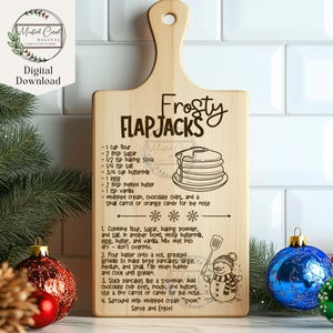 May include: A wooden cutting board with a recipe for "Frosty Flapjacks" engraved on it. The recipe includes ingredients like flour, sugar, baking soda, and buttermilk. The board also features a snowman illustration and a stack of pancakes.