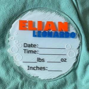 May include: A white circular acrylic plaque with a blue and orange name "Elian Leonardo" and spaces to record the date, time, weight in pounds and ounces, and length in inches.