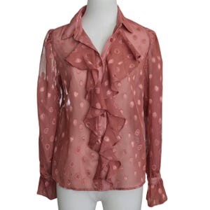May include: A sheer, dusty rose-colored blouse with a ruffled front and small, circular polka dots. The long-sleeved top features a collared neckline and button-down closure. The cuffs are flared.
