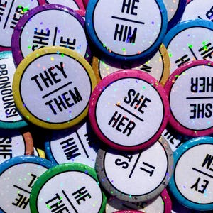 May include: A collection of circular pin-back buttons featuring various pronoun sets printed in black text on white backgrounds. Each button has a colorful border and a holographic glitter finish that reflects light across the surface.