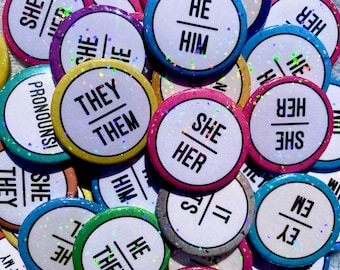 May include: A collection of circular pin-back buttons featuring various pronoun sets printed in black text on white backgrounds. Each button has a colorful border and a holographic glitter finish that reflects light across the surface.