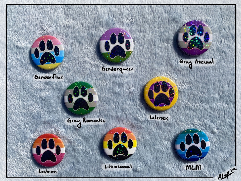 May include: A collection of round, colorful buttons with paw print designs. Each button features a different pride flag color scheme and a label, including "Genderflux," "Genderqueer," "Gray Asexual," "Gray Romantic," "Intersex," "Lesbian," "Lithiosexual," and "MLM."