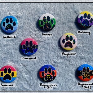 May include: A collection of nine round buttons with paw print designs, each featuring a different color scheme and text label. The buttons represent various LGBTQ+ identities, including "Neptunic," "Omnisexual," and "Nonbinary."