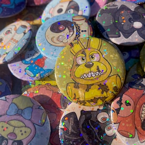 May include: A collection of colorful, glittery, round buttons featuring various cartoon characters. The buttons are all different sizes and have a holographic finish.
