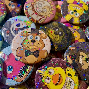 Peut inclure: Une collection de boutons brillants et colorés représentant des personnages de la franchise de jeux vidéo populaire Five Nights at Freddy's. Les boutons présentent divers personnages, dont Freddy Fazbear, Chica la Poule, Foxy le Renard Pirate et Bonnie le Lapin. Les boutons sont tous ronds et ont une finition brillante et holographique.