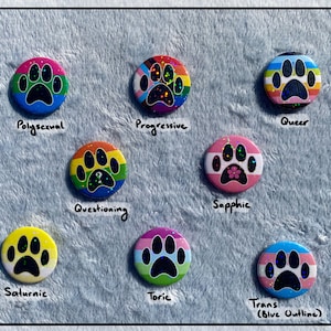 May include: A collection of round, colorful buttons featuring paw prints and various LGBTQ+ pride flags. Each button has a different design, including the words "Pollysexual", "Progressive", "Queer", "Questioning", "Sapphic", "Saturnic", "Toric", and "Trans (Blue Outline)".