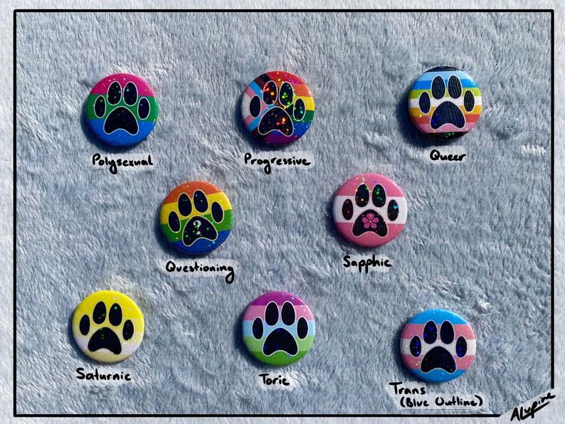 May include: A collection of round, colorful buttons featuring paw prints and various LGBTQ+ pride flags. Each button has a different design, including the words "Pollysexual", "Progressive", "Queer", "Questioning", "Sapphic", "Saturnic", "Toric", and "Trans (Blue Outline)".