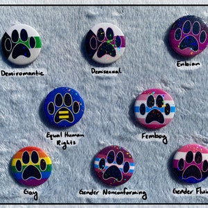 May include: A collection of round, colorful buttons with paw print designs, each representing different identities. The buttons feature text such as "Demiroamantic", "Demisexual", "Enbian", "Equal Human Rights", "Femboy", "Gay", "Gender Nonconforming", and "Gender Fluid".
