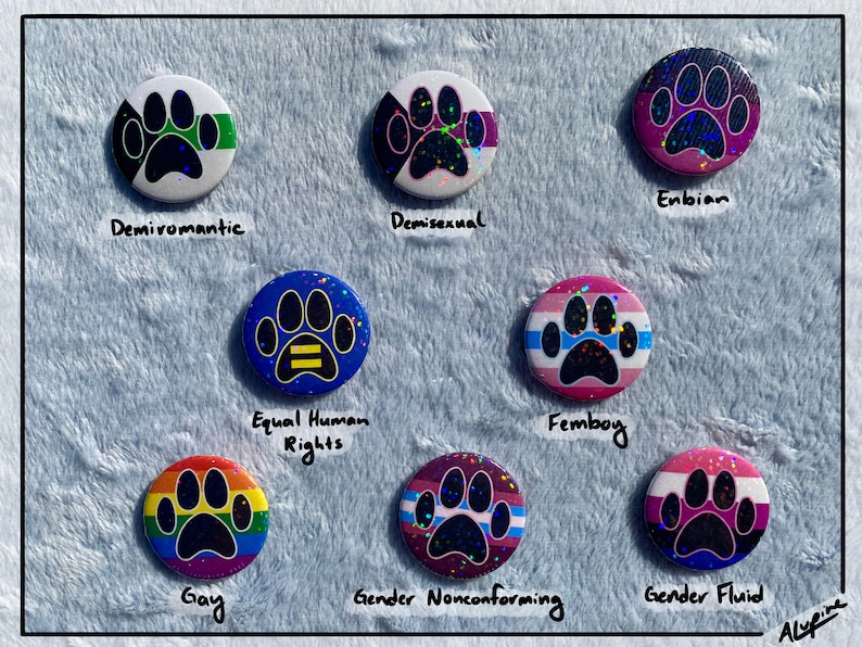 May include: A collection of round, colorful buttons with paw print designs, each representing different identities. The buttons feature text such as "Demiroamantic", "Demisexual", "Enbian", "Equal Human Rights", "Femboy", "Gay", "Gender Nonconforming", and "Gender Fluid".