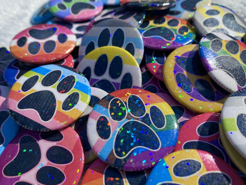 May include: A collection of colorful, round buttons featuring a paw print design. The buttons display various rainbow and pride flag patterns, with a glittery, iridescent finish. The buttons are a vibrant mix of colors, including pink, blue, yellow, and purple.