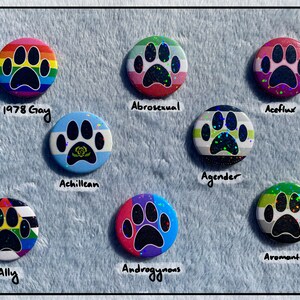 May include: A collection of round, colorful buttons featuring paw prints. Each button represents a different LGBTQ+ identity, with labels such as "1978 Gay," "Abrosexual," and "Aromantic." The buttons have a holographic, glittery effect.