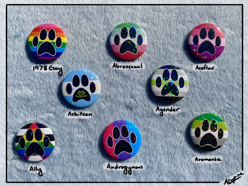 May include: A collection of round, colorful buttons featuring paw prints. Each button represents a different LGBTQ+ identity, with labels such as "1978 Gay," "Abrosexual," and "Aromantic." The buttons have a holographic, glittery effect.