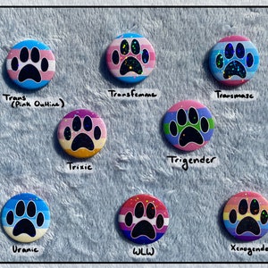 May include: A collection of nine round buttons with paw print designs. Each button features a different color scheme and text, including "Trans," "Transfemme," "Transmasc," "Trixie," "Trigender," "Uranic," "WLL," and "Xenogender."