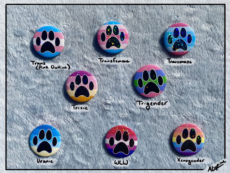May include: A collection of nine round buttons with paw print designs. Each button features a different color scheme and text, including "Trans," "Transfemme," "Transmasc," "Trixie," "Trigender," "Uranic," "WLL," and "Xenogender."