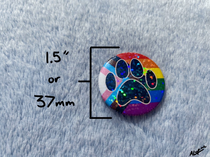 May include: A round button with a rainbow paw print design, approximately 1.5 inches or 37mm in diameter. The button features a rainbow background and a glittery paw print. The button has a transgender pride flag design on the left side.