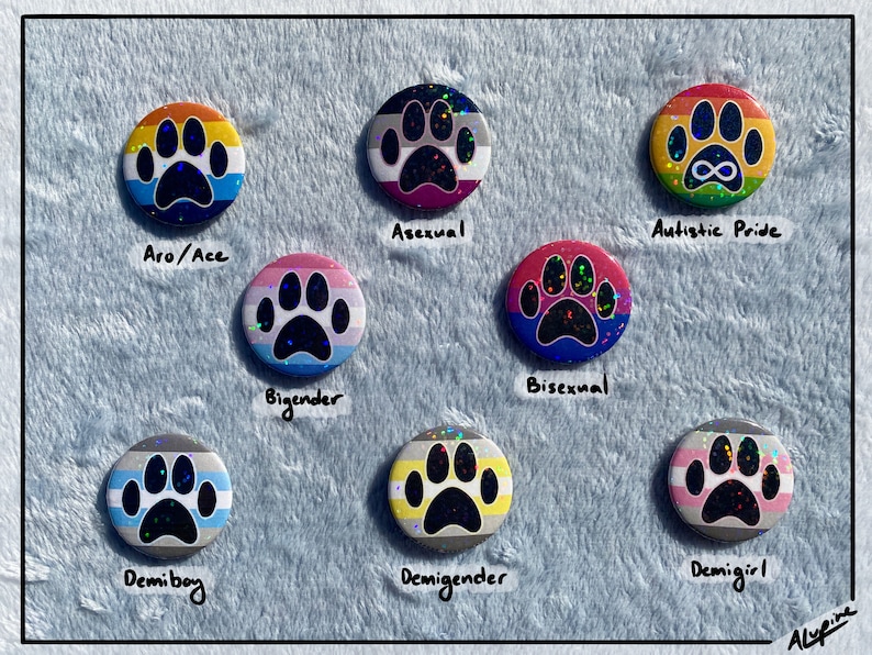 May include: A collection of round, colorful pride buttons featuring a paw print design. Each button displays a different flag design, including Aro/Ace, Asexual, Autistic Pride, Bigender, Bisexual, Demiboy, Demigender, and Demigirl.