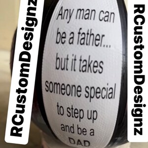 Op de afbeelding: Een zwart-witte afbeelding van een rond object met de tekst "Any man can be a father... but it takes someone special to step up and be a DAD" erop gedrukt. De tekst "RCustomDesignz" is links en rechts op het object gedrukt.