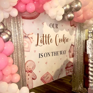 May include: A white backdrop with pink and silver balloons, a gingerbread man, and the text "Our Little Cookie Is On The Way".