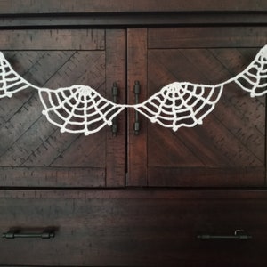 May include: A white crocheted spiderweb garland with five web designs hanging on a dark brown wooden cabinet.