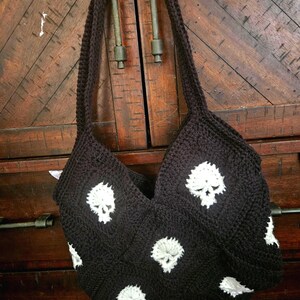 May include: A black crochet tote bag featuring white skull motifs. The bag has a long strap and is displayed against a wooden background. The skull design is repeated across the front and sides of the bag.