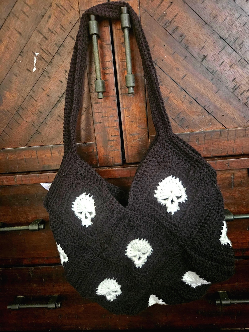 May include: A black crochet tote bag featuring white skull motifs. The bag has a long strap and is displayed against a wooden background. The skull design is repeated across the front and sides of the bag.