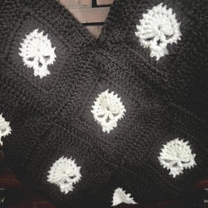 May include: Black and white crocheted blanket featuring a repeating skull pattern. The skulls are white and the background is black. The blanket is made of a thick, textured yarn, and is folded over.