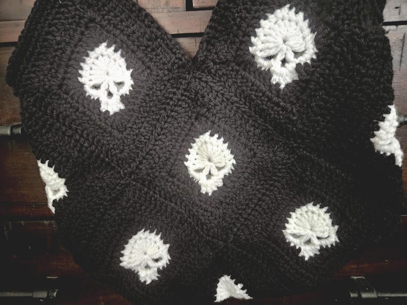 May include: Black and white crocheted blanket featuring a repeating skull pattern. The skulls are white and the background is black. The blanket is made of a thick, textured yarn, and is folded over.