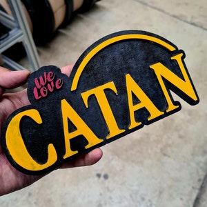 May include: A black wooden sign with the word "CATAN" in yellow, and the words "We Love" in pink. The sign has a curved top and is held in a hand.