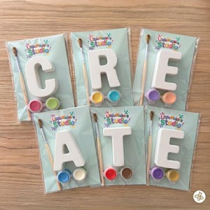 Könnte beinhalten: Sechs einzelne Bastelsets in durchsichtiger Plastikverpackung, die jeweils einen weißen Buchstaben, einen Holzpinsel und zwei kleine Farbtöpfe enthalten. Die Buchstaben bilden das Wort CREATE und tragen das Little Makers Studio Logo.
